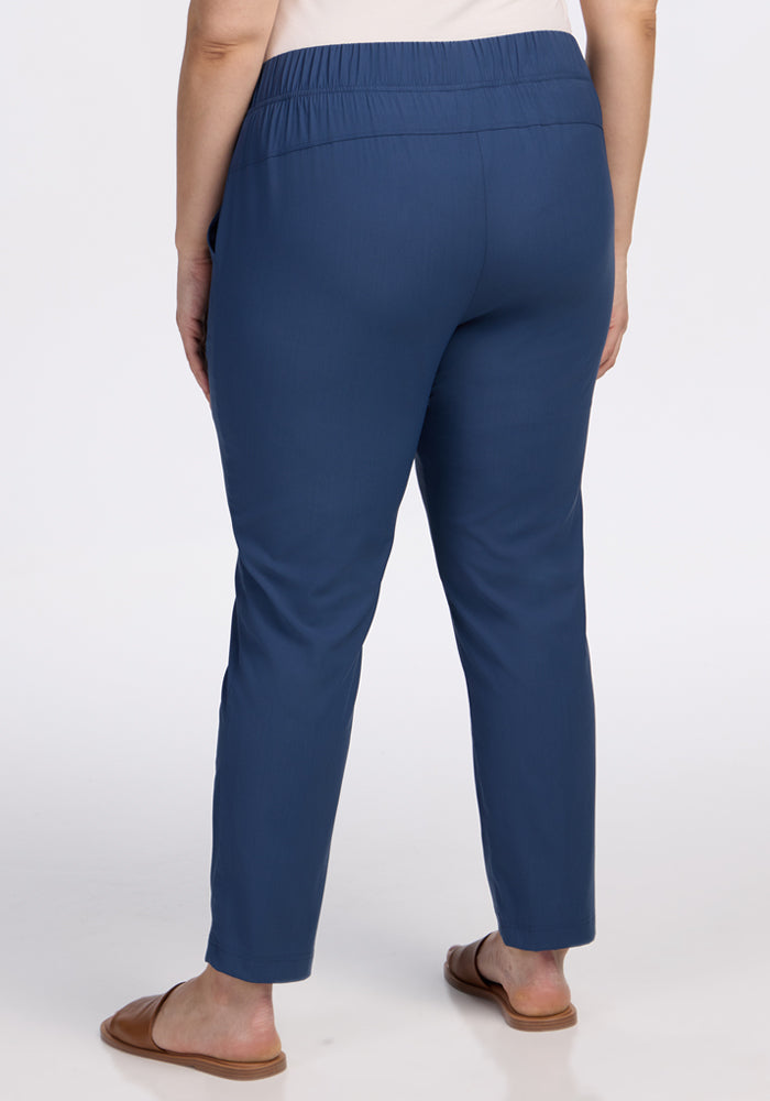 A person is shown from the waist down, wearing Woolx Nicole Ankle Pants in Vintage Indigo with an elastic waistband and brown flat sandals, standing against a plain white background.