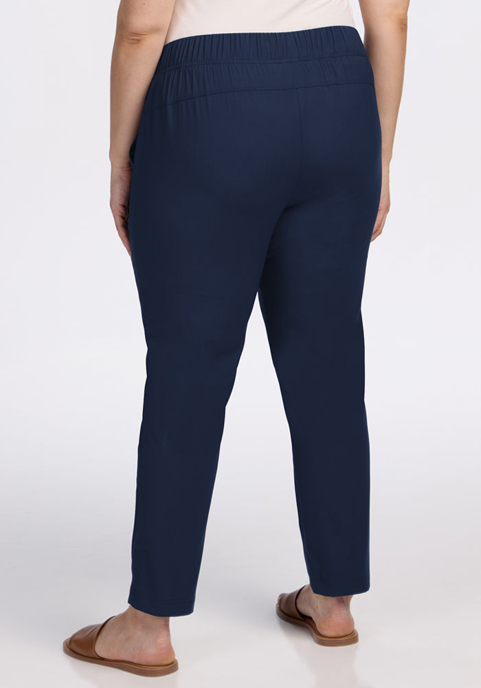 A person faces away from the camera, wearing Woolx Nicole Ankle Pants in Deep Navy with an elastic waistband, a light top, and brown slide sandals, set against a plain white background.