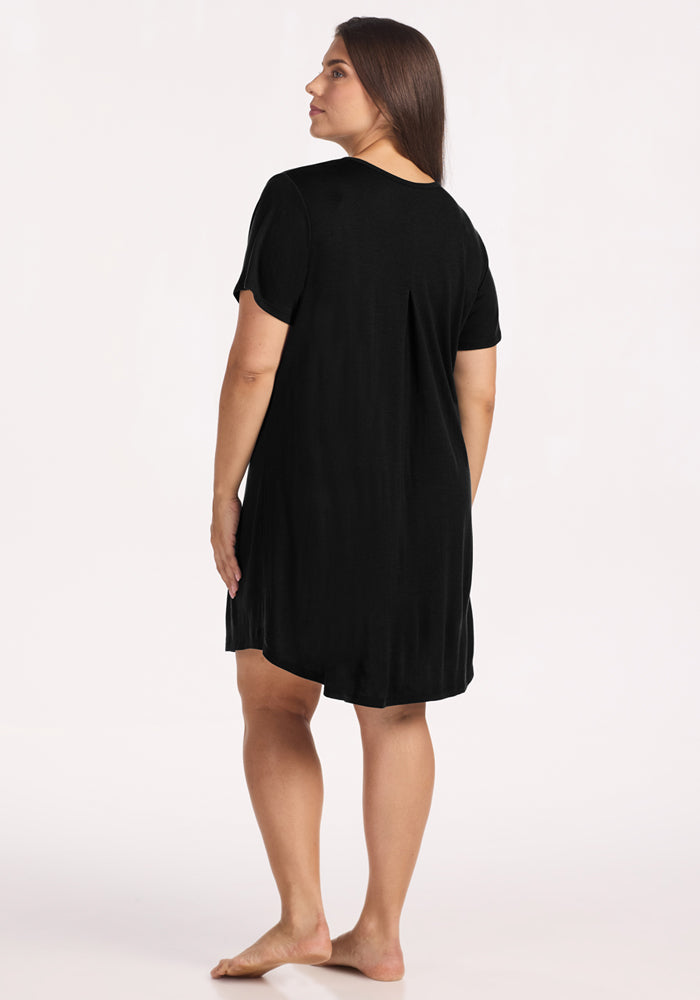 A woman with long brown hair stands barefoot, facing away from the camera, wearing the loose-fitting Woolx Desi Nightgown in black against a plain light background.
