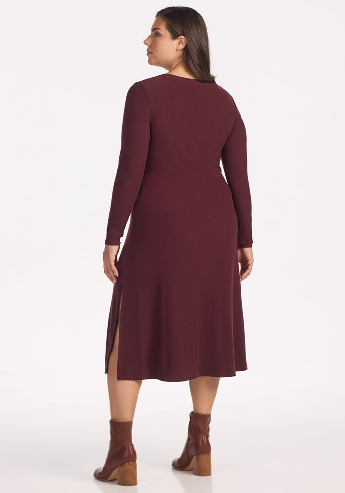 A woman faces away, wearing the Woolx Caroline Ribbed Twirl Dress in Toasted Garnet with long sleeves, knee-length hem and side slits, paired with brown ankle boots against a plain white background.