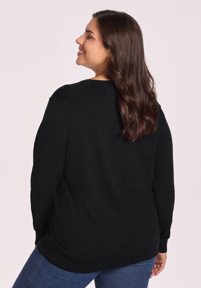 A woman with long brown hair smiles over her shoulder while wearing the Woolx Francesca Sweater in black and blue jeans, standing against a light background.