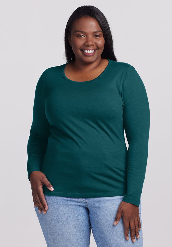 A woman with long dark hair smiles while wearing the Woolx Remi Long Sleeve T-Shirt in Riverbank Green and light blue jeans, standing against a plain light gray background with one hand by her side. 