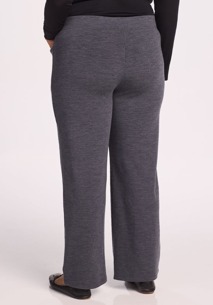 A person wearing Woolx Ellie Wide Leg Pants in Cinder Melange and black flats stands with their back to the camera. The cozy, straight-leg Merino wool pants are styled with a long-sleeve black top.