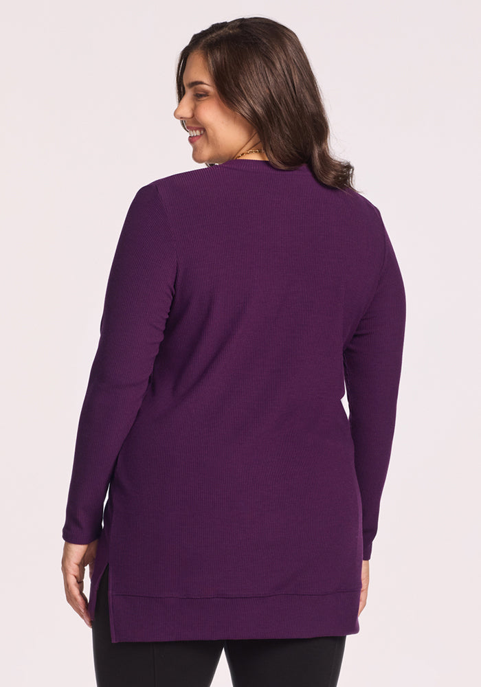 A woman with medium-length brown hair stands with her back to the camera, wearing the Woolx Evie Ribbed Tunic in Majestic Purple and black pants against a plain light background.