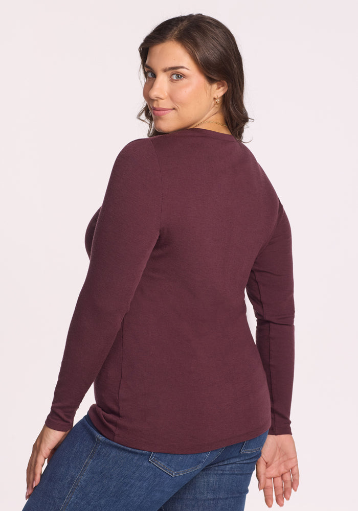 A woman with long brown hair stands against a plain light background, looking over her shoulder in the Woolx Reese Ribbed Henley - Chocolate Fig and blue jeans.
