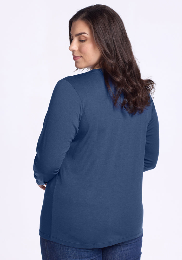 A woman with long brown hair, wearing the Woolx Millie Long Sleeve in Starry Night and blue jeans, stands facing away from the camera and looks over her shoulder with a neutral expression.