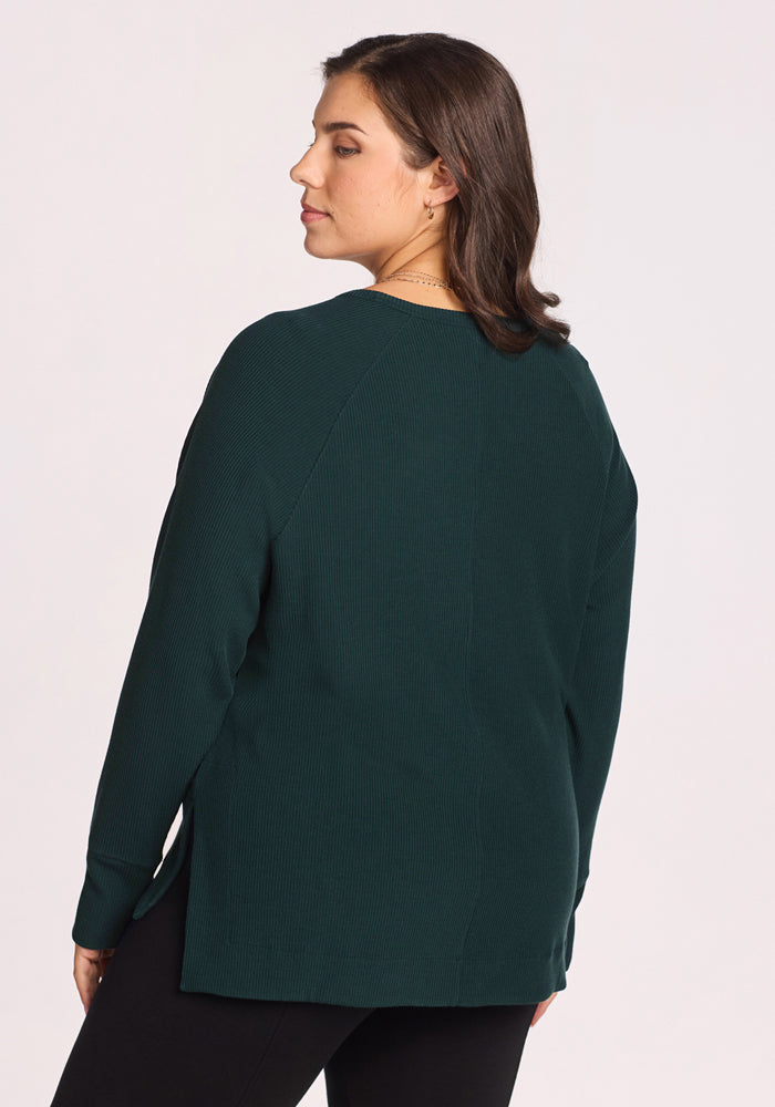 A woman with shoulder-length brown hair stands facing away from the camera, wearing the Woolx Ainsley Sweater in Deep Timber and black pants against a plain light background.