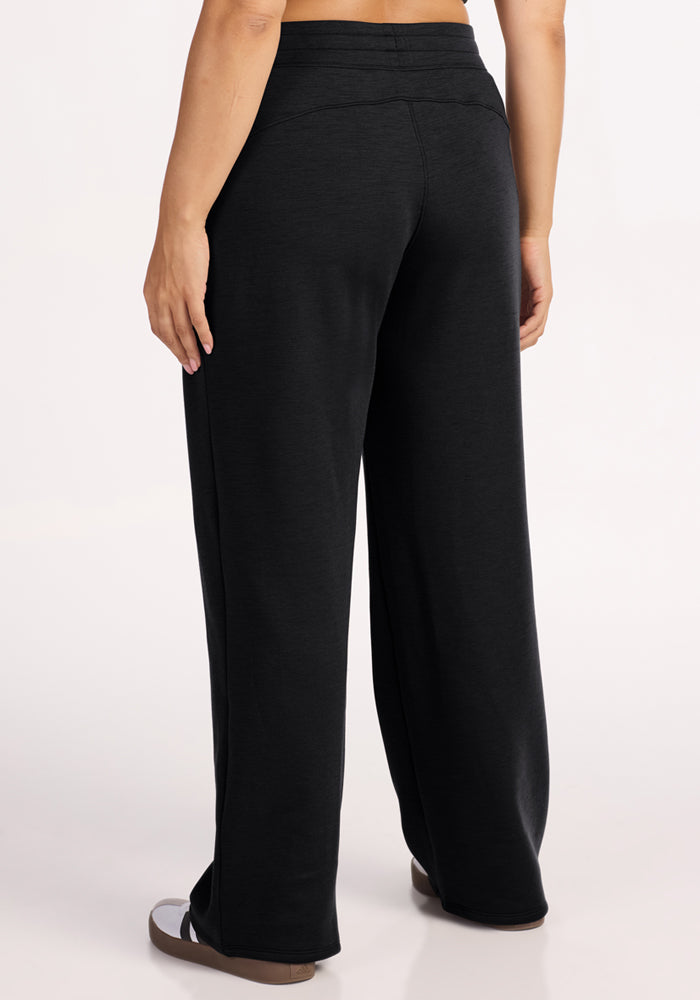 A person wears Woolx Bree Lounge Pants in black and sandals, standing on a plain light background. Only the lower half is visible.