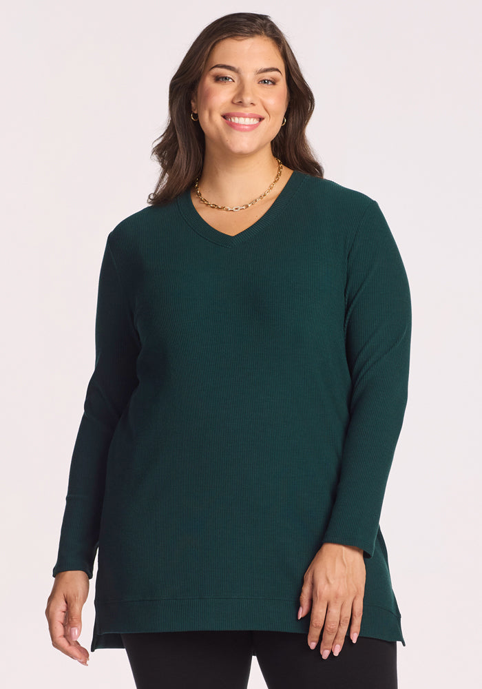 A woman with long brown hair, wearing the Woolx Evie Ribbed Tunic in Deep Timber and black pants, stands smiling against a plain light background. 