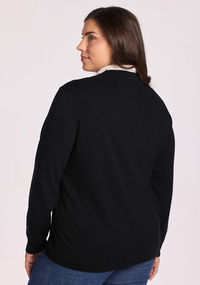 A woman with long brown hair stands facing away, wearing the Woolx Gia Sweater in black over a white collared shirt and blue jeans. A soft pink background highlights her chic, relaxed style.