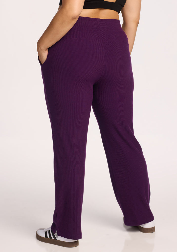 A person is seen from the back wearing Woolx Luca Ribbed Pants in Majestic Purple with pockets and a wide leg, paired with black-and-white sneakers. The relaxed-fit pants stand out against a plain, light background.