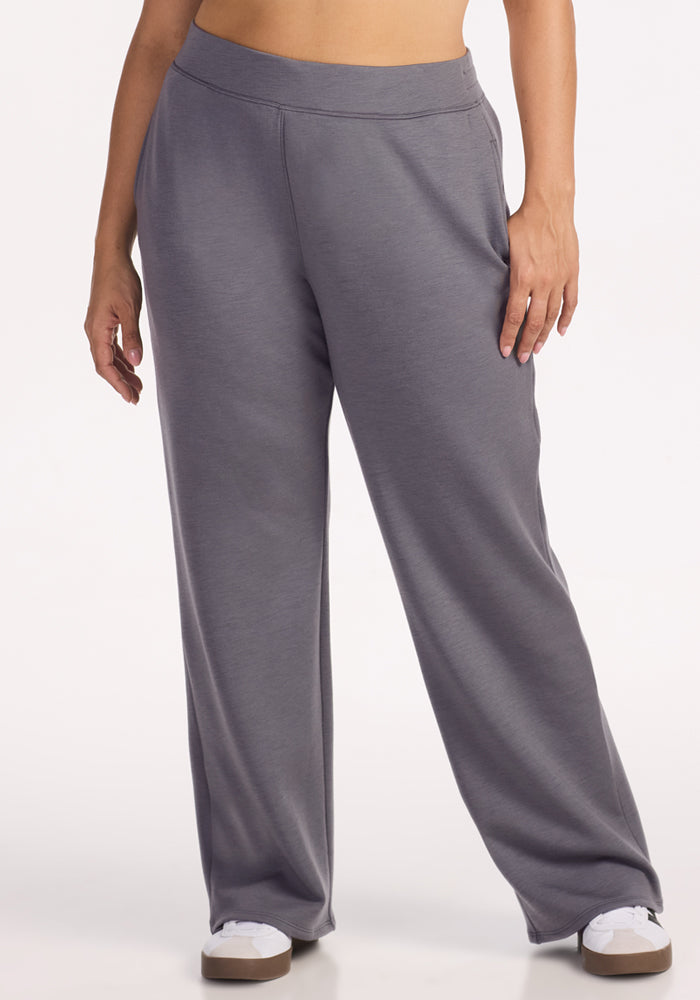 A person wears Woolx Bree Lounge Pants in Arctic Dusk, featuring a relaxed fit, elastic waistband, and wide legs. Standing with one hand on their thigh, they pair the gray pants with white sneakers against a plain white background.