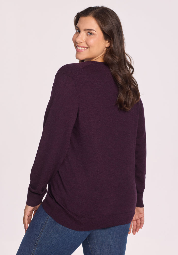 A woman with long brown hair smiles over her shoulder, wearing the Woolx Francesca Sweater in Deep Plum and blue jeans, standing against a plain light background.