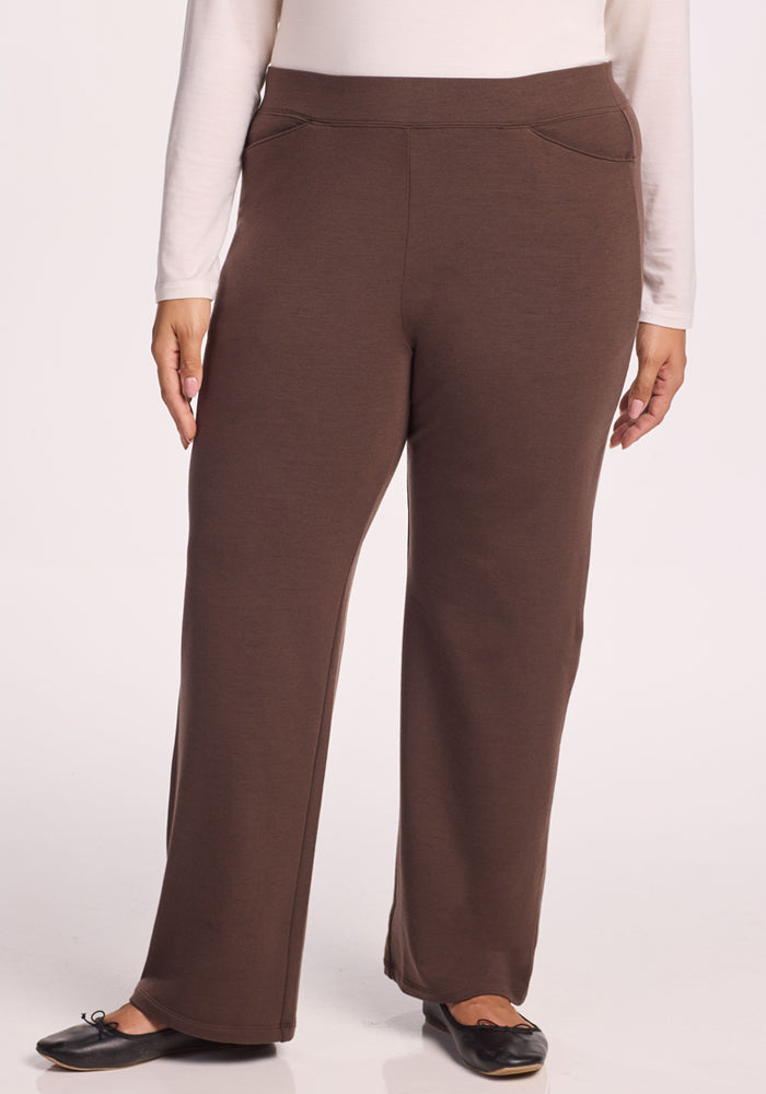 A person models the Woolx Ellie Wide Leg Pants in Bracken, featuring a wide waistband and straight-leg Merino wool design, paired with a white long-sleeve top. Only the lower half of their body is visible against a plain white background. 