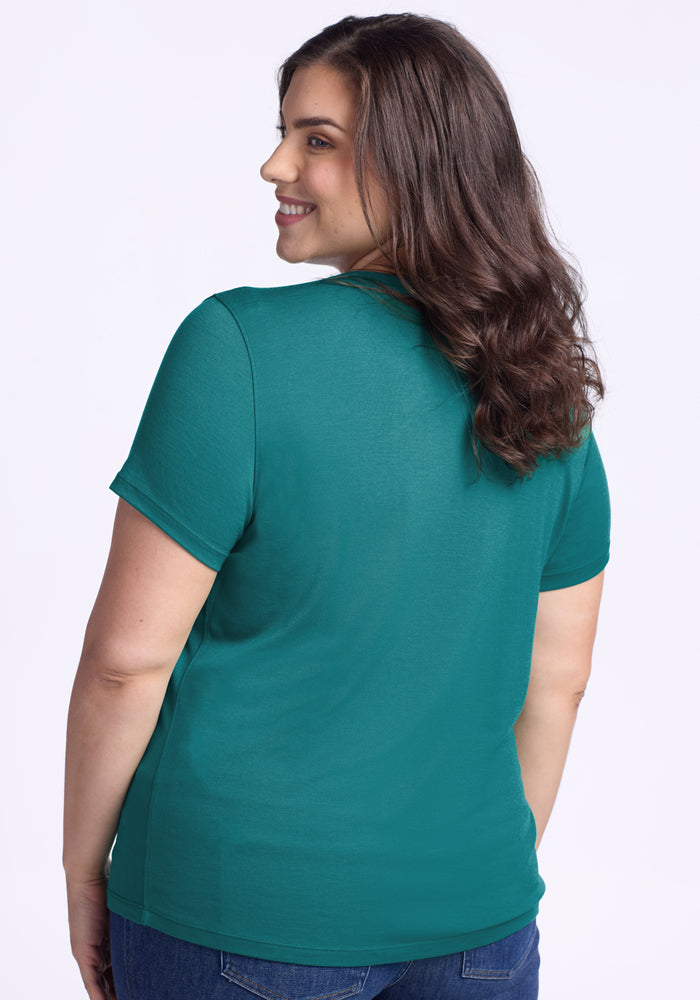 A woman with wavy brown hair smiles over her shoulder in a Woolx Liza Crew Neck Tee – Surfside and blue jeans, standing against a plain light background.