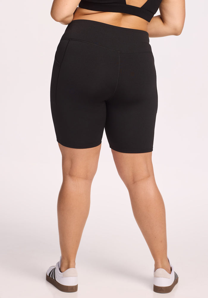 A person faces away, wearing a black sports bra, Woolx Merino Flex® Dani Bike Shorts in black, and white sneakers with brown soles against a light background.
