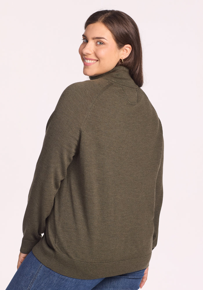 A woman with long brown hair, wearing the Woolx Collins Half Zip Sweater in Pine Needle and blue jeans, stands turned slightly to the side and smiles over her shoulder against a plain light background.