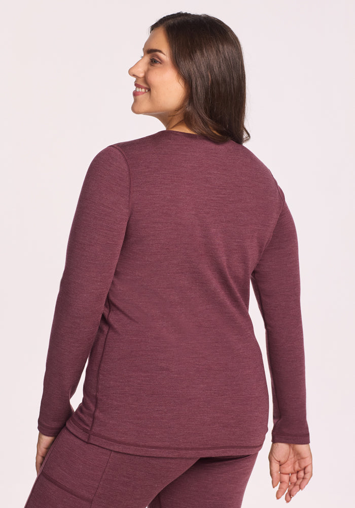 A woman with long brown hair smiles over her shoulder, wearing the Woolx Elsa Long Sleeve in Grape Harvest Melange. She stands against a light background, highlighting its cozy, moisture-wicking merino wool comfort.
