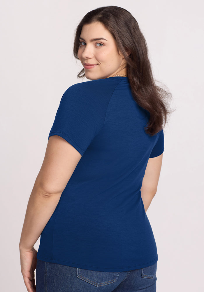 A woman with long brown hair models the Woolx Mia V Neck in Estate Blue, paired with blue jeans. She stands with her back to the camera, smiling over her shoulder against a plain light background.
