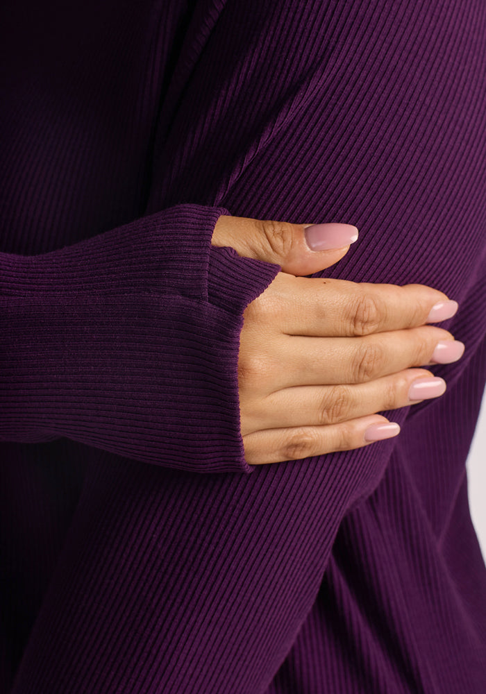 A person with manicured light pink nails rests their hand on their arm, wearing the Woolx Ainsley Sweater in Majestic Purple—a ribbed Merino wool piece with a thumb hole for temperature-regulating comfort and travel-ready style.