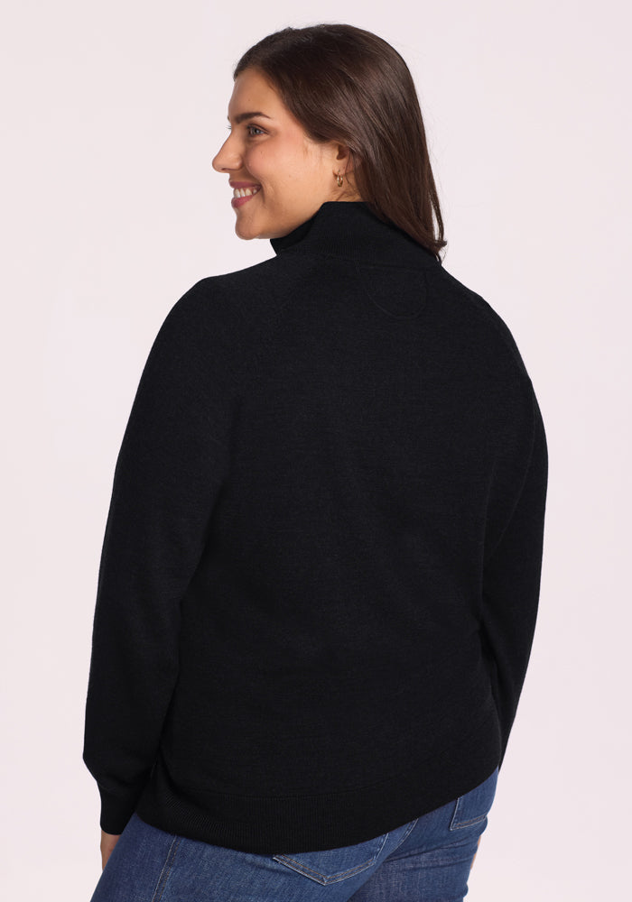 A woman with long brown hair smiles while wearing the Woolx Collins Half-Zip Sweater in black and blue jeans, photographed from the back and turned slightly to the side against a plain light background.