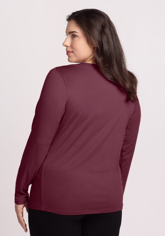 A woman with long brown hair stands against a light background, wearing the Woolx Millie Long Sleeve in Grape Harvest, a Merino wool top known for its temperature-regulating properties. Her head is turned slightly to the side.
