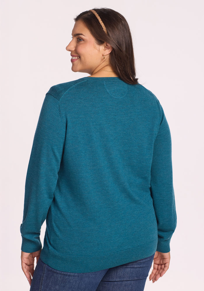 A woman with long dark hair, wearing the Woolx Francesca Sweater in Harbor Teal and blue jeans, stands facing away and smiles over her shoulder.