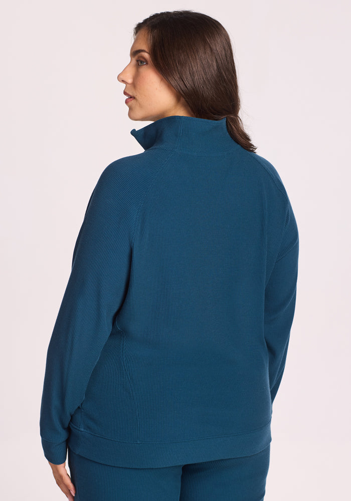 A woman with long brown hair wears the Woolx Quinn Ribbed 1/4 Zip in Majolica Blue, paired with matching pants. She stands with her back to the camera, head slightly left, against a light background.