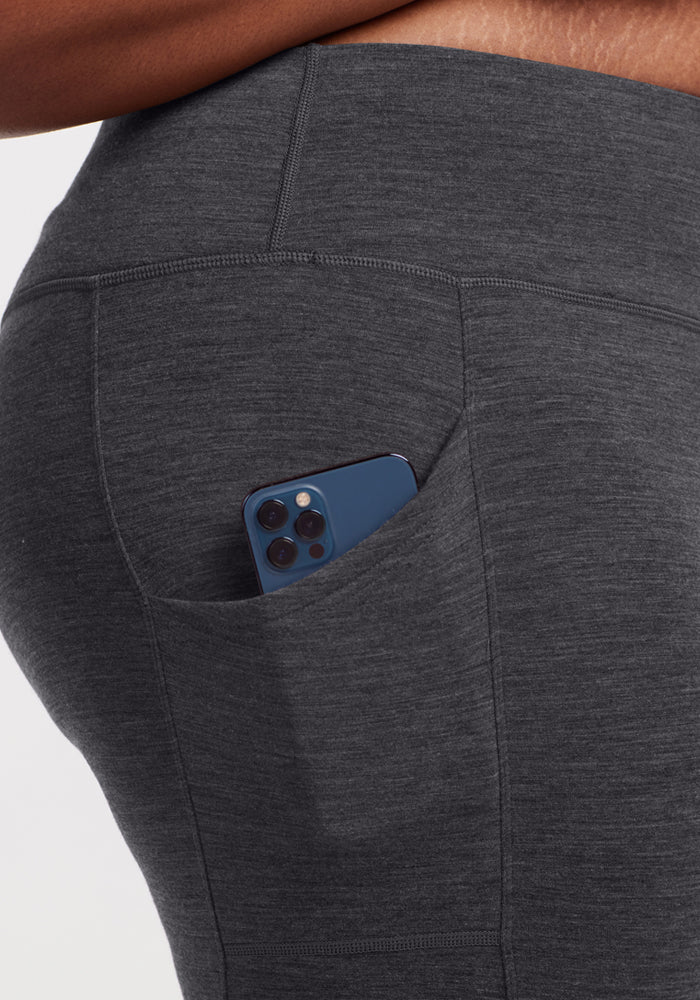 Close-up of a person wearing Woolx Piper Pocket Leggings in Cinder Melange, featuring moisture-wicking fabric and a blue smartphone partially tucked into the side pocket.
