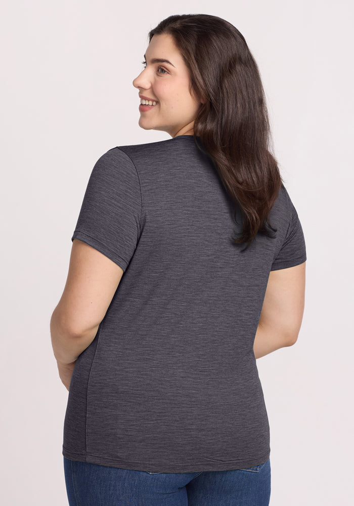 A woman with long dark hair wears the Woolx Liza Crew Neck Tee in Cinder Melange and blue jeans, standing with her back to the camera and smiling over her shoulder against a light background.