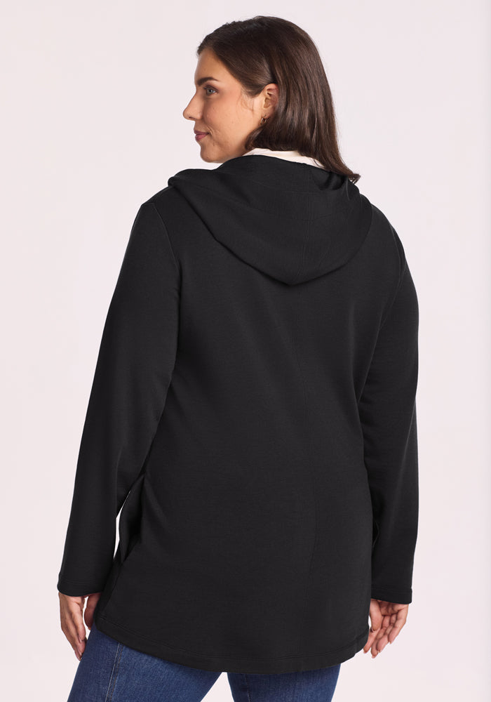 A woman with medium-length brown hair is shown from the back, wearing the Woolx Edna Hooded Cardigan in black with a white lining and blue jeans, standing against a plain light background.