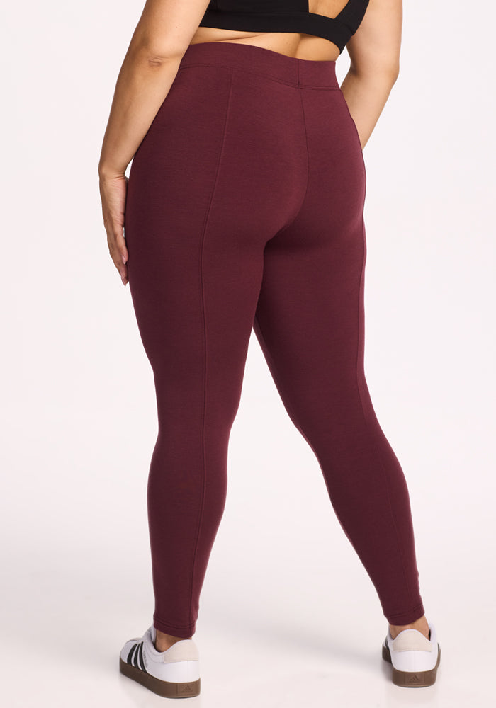 A person wearing a black crop top, Woolx Stella Leggings in Toasted Garnet, and white sneakers stands with their back to the camera against a plain white background.