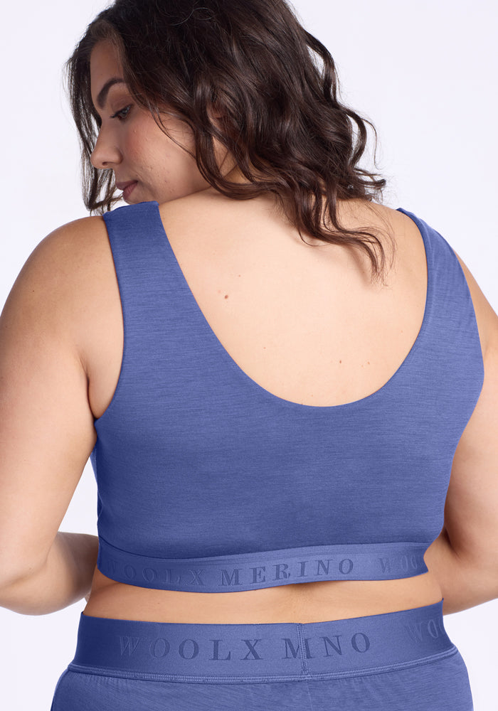 A woman with wavy brown hair is shown from the back wearing the Woolx Aubrey Lounge Bra in Cloudy Bay, paired with matching underwear. The bra features a scoop back and a waistband that reads "WOOLX MERINO.