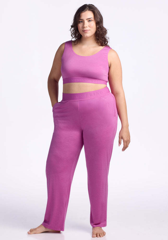 A woman with wavy brown hair models the Woolx Aubrey Lounge Bra in Wild Blossom, paired with high-waisted lounge pants. She stands barefoot against a plain white background, one hand in her pocket and wearing a relaxed expression.