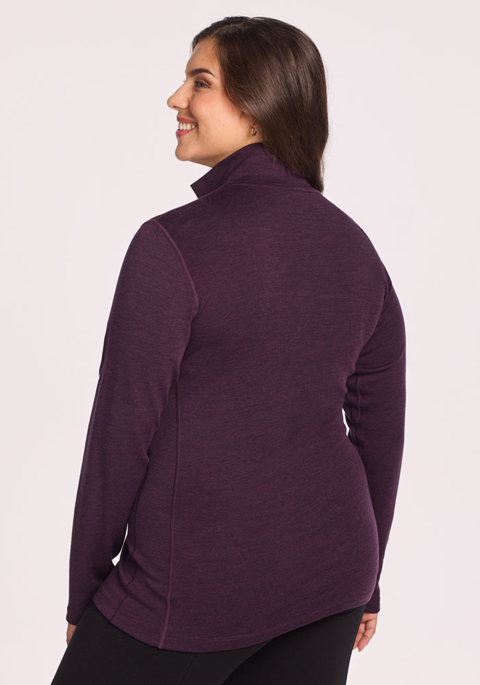 A woman with long brown hair smiles, facing away from the camera, wearing the Woolx Brooke 1/4 Zip in Deep Plum and black pants—ideal hiking performance attire—against a plain light background.