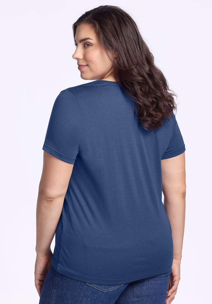 A woman with long brown hair, wearing the Woolx Junie V Neck - Starry Night and jeans, stands facing away but glances over her shoulder with a slight smile. The background is plain and light-colored.