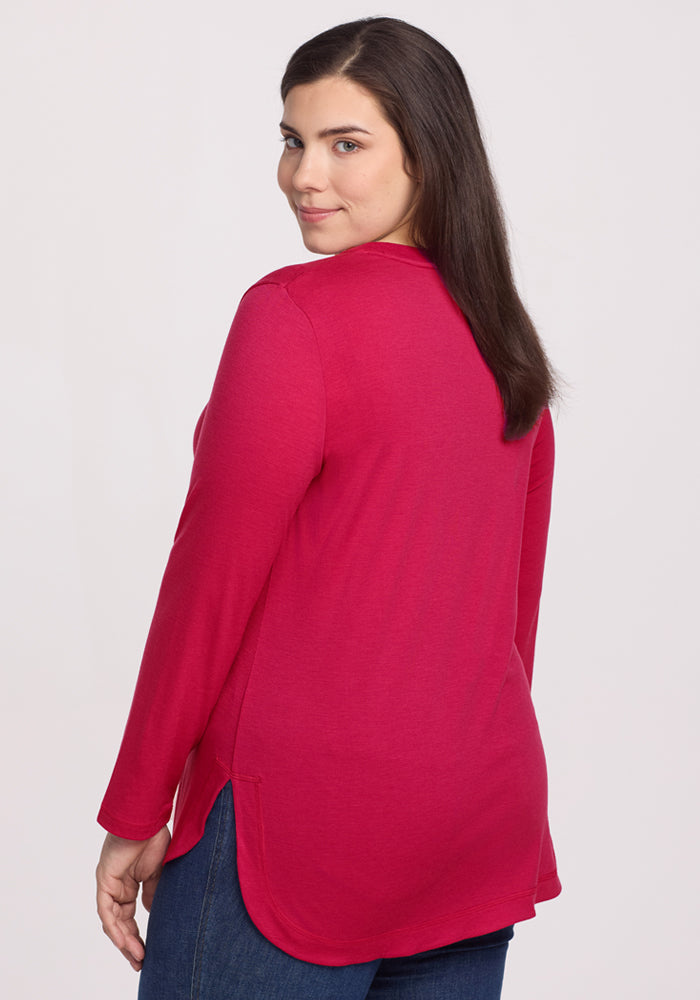 A woman with long brown hair wears the Woolx Hazel Tunic in Poppy Spark and blue jeans, standing against a light background. She faces away but glances back over her shoulder with a slight smile.