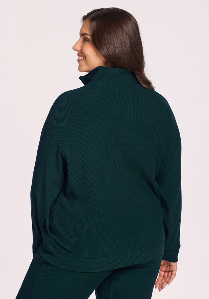 A woman with long brown hair, wearing the Woolx Quinn Ribbed 1/4 Zip in Deep Timber and matching pants, stands facing away and smiles over her shoulder against a plain light background.