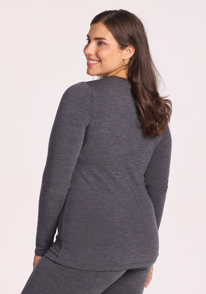 A woman with long brown hair, wearing the Woolx Sadie Baselayer Top in Cinder Melange, stands turned slightly away and smiles over her shoulder against a light background.