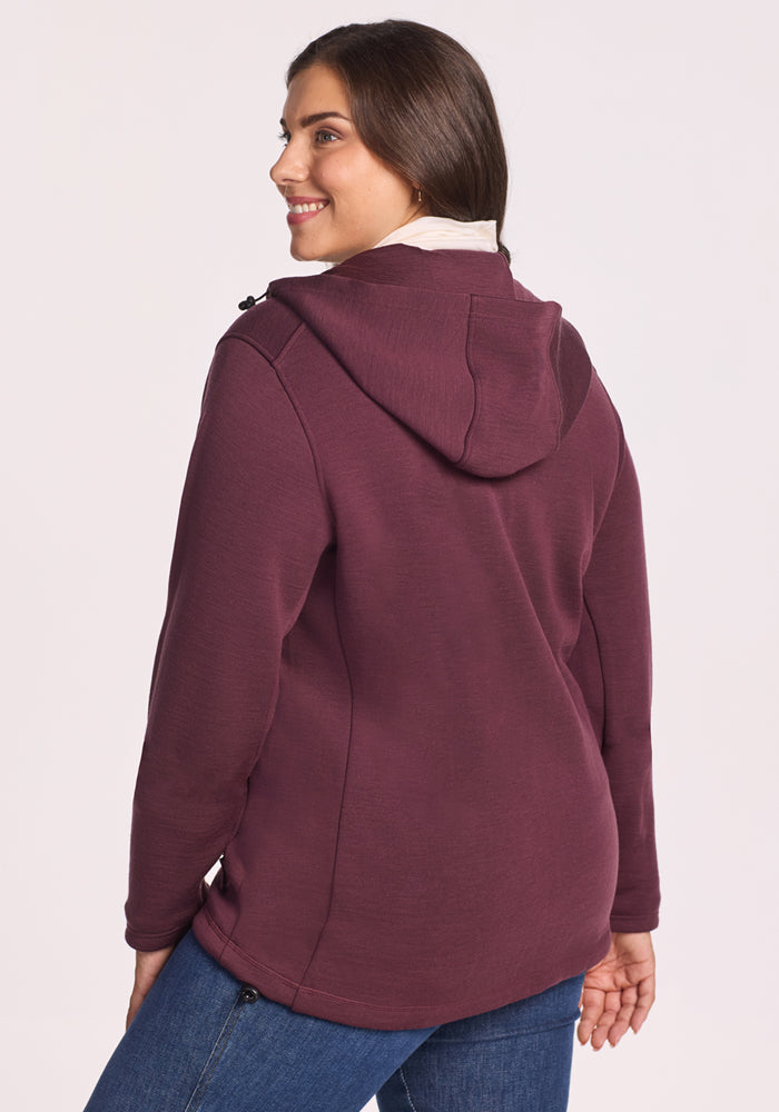 A woman with long brown hair, wearing the Woolx Cubby Hooded Sweatshirt in Grape Harvest and blue jeans, stands smiling with her back to the camera against a plain light background.