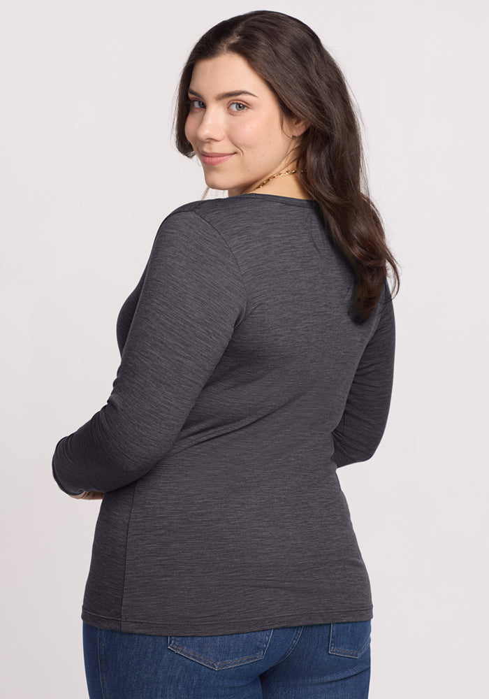 A woman with long brown hair, wearing the Woolx Layla V Neck Top in Cinder Melange and blue jeans, stands turned slightly away from the camera and looks back over her shoulder with a slight smile.