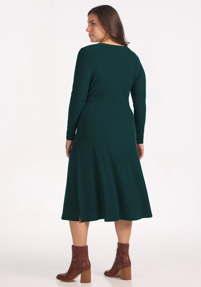 A woman faces away, wearing the Woolx Caroline Ribbed Twirl Dress in Deep Timber, featuring long sleeves and a slight flare with side slit, paired with brown ankle boots against a plain white background.