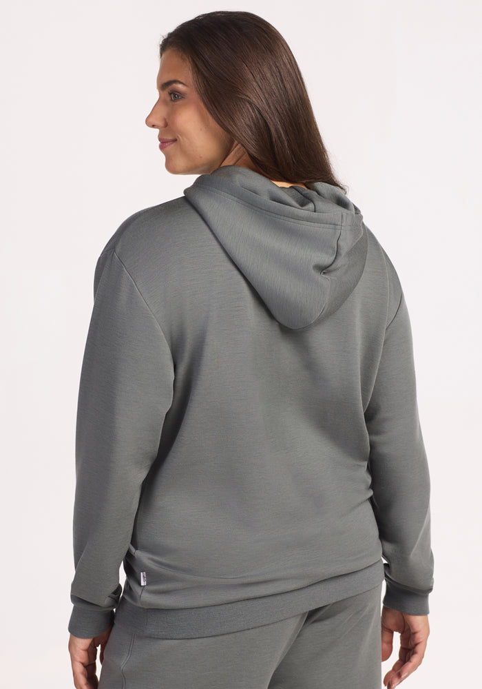 A woman with long brown hair wears the Woolx Avery Hoodie in Sedona Sage, paired with matching pants, standing against a plain light background and looking over her shoulder.