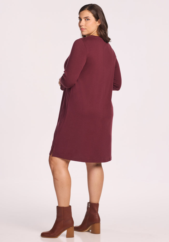 A woman with medium dark hair stands facing away, wearing the Woolx Rissa A Line Dress in Toasted Garnet—knee-length merino wool, ideal for fall and winter layering—and brown block-heeled ankle boots against a plain light background.