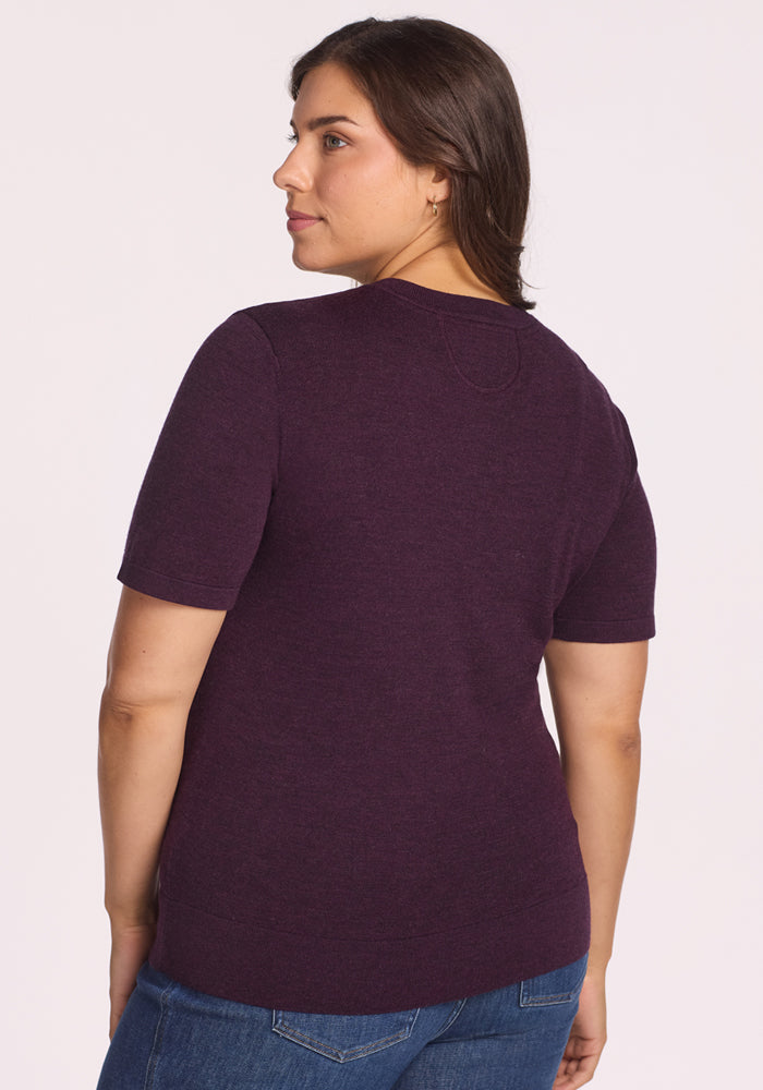 A woman with medium-length brown hair is shown from the back, wearing the Woolx Anna Crew Sweater in Deep Plum with blue jeans, standing against a plain light background.