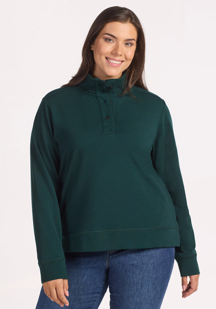 A woman with long brown hair, wearing the Woolx Annie 3 Button Pullover in Deep Timber and blue jeans, smiles against a plain white background. 