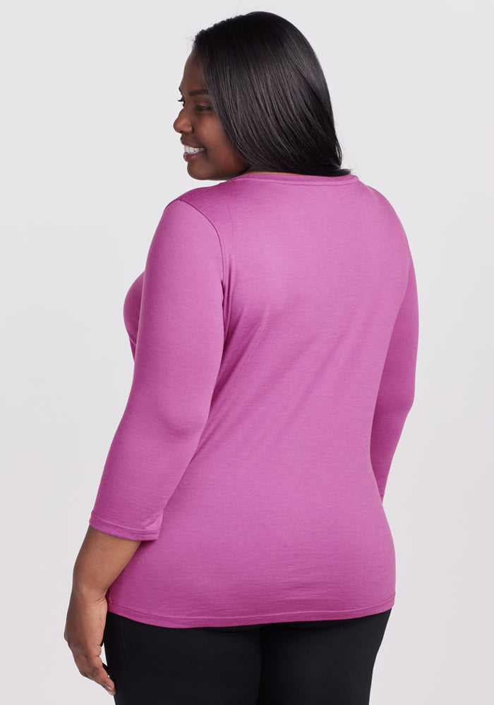 A woman with long dark hair is shown from the back, wearing the Woolx Jenny 3/4 Sleeve Crew Neck in Purple Wine and black pants. She stands against a plain light background, smiling slightly.