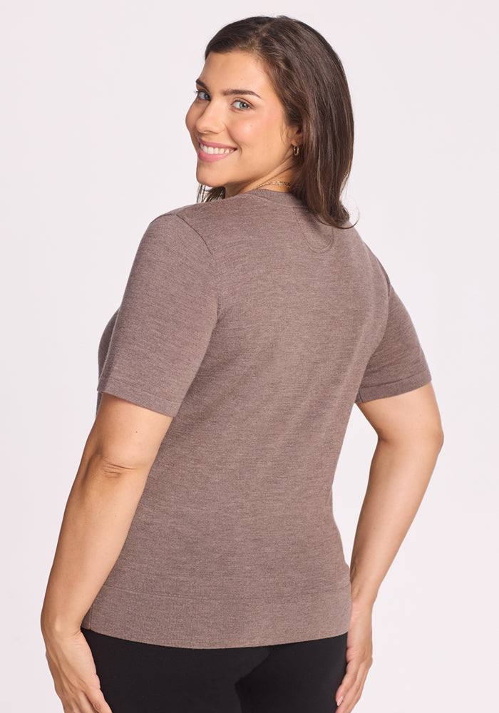 A woman with long brown hair smiles over her shoulder, wearing the Woolx Anna Crew Sweater in Simply Taupe and black pants, standing against a plain light background.