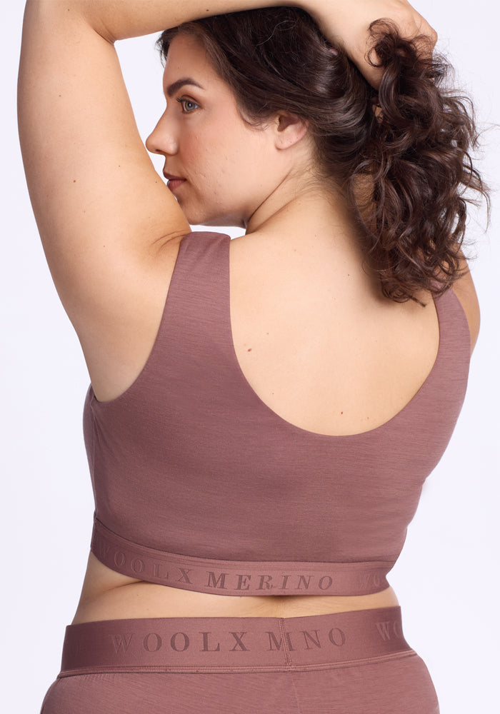A woman with curly brown hair poses with one arm raised, wearing the Woolx Aubrey Lounge Bra in Nutmeg and matching high-waisted underwear. The bra back reads "MERINO," and the underwear has a Woolx logo waistband.