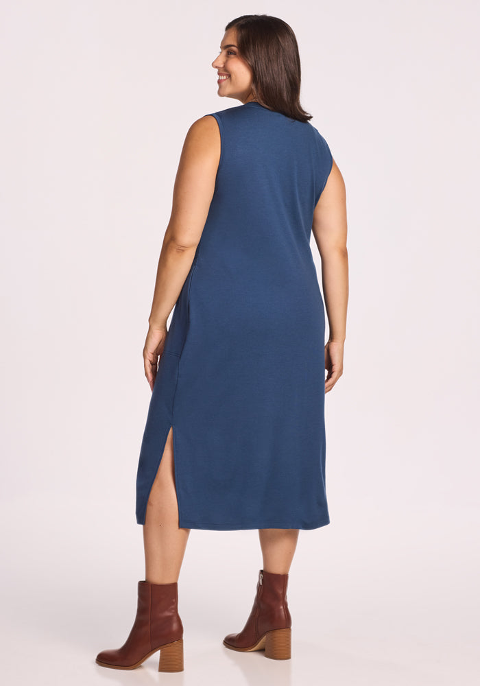 A woman models the Woolx Cassie Dress - Starry Night, a sleeveless, knee-length blue Merino wool dress with a back and side slit. She faces away, smiling with straight brown hair, brown ankle boots, and a plain white background.
